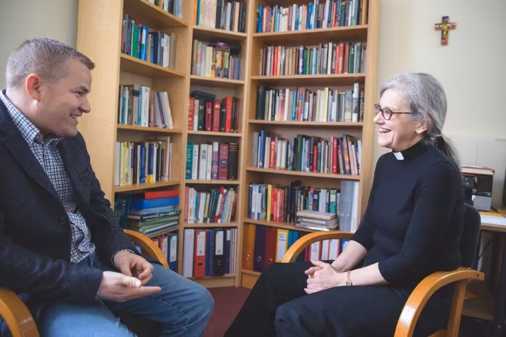 Two people sitting talking to each other in front of  well-stocked bookcases