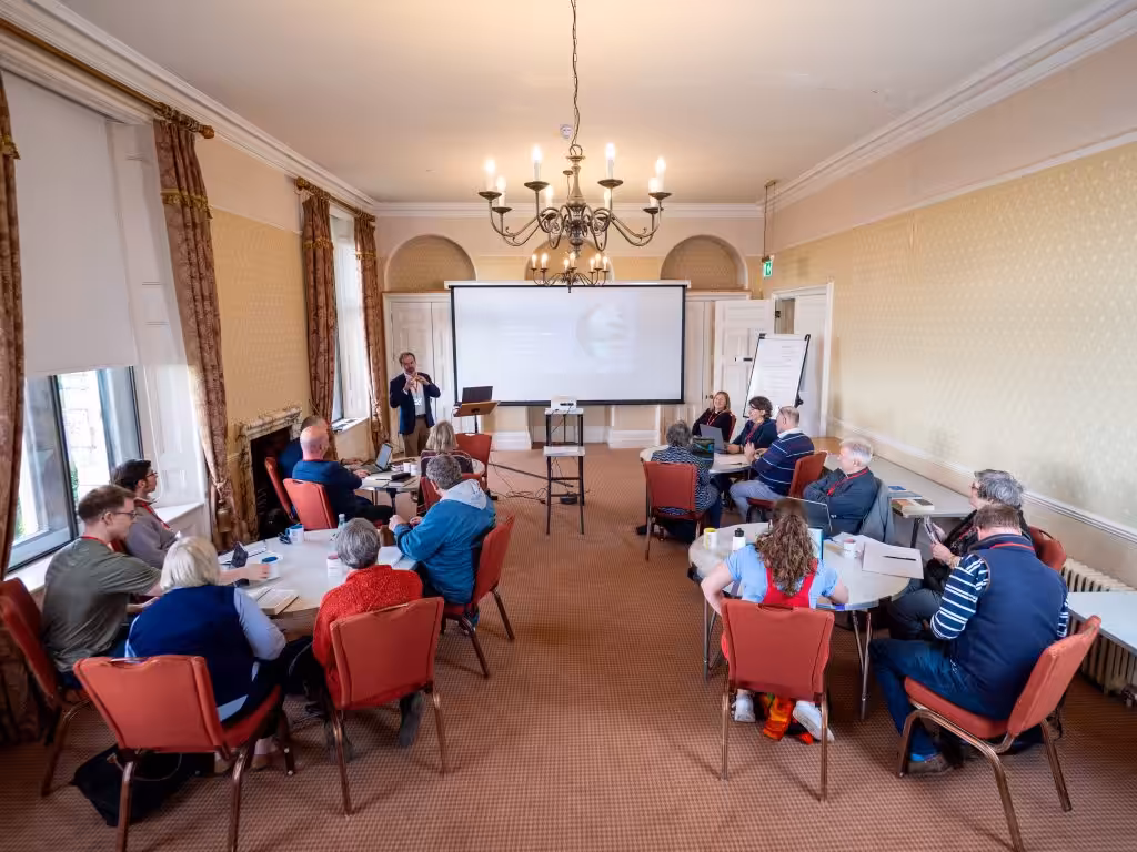 A room with people sitting in rows watching a person speak with a whiteboard behind them